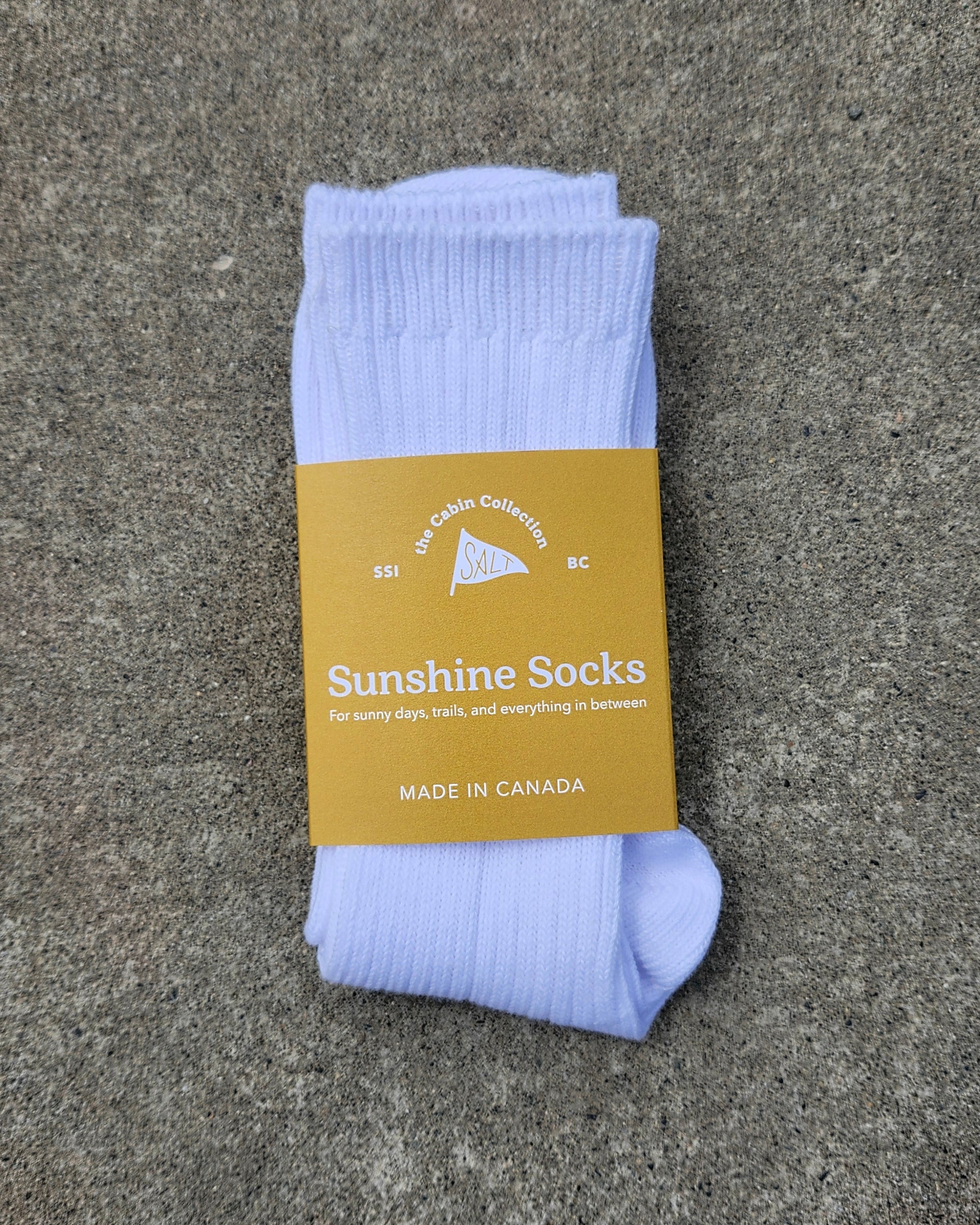 A pair of light white ribbed socks folded with a mustard yellow paper label that reads 'Sunshine Socks', placed on a textured gray concrete surface.