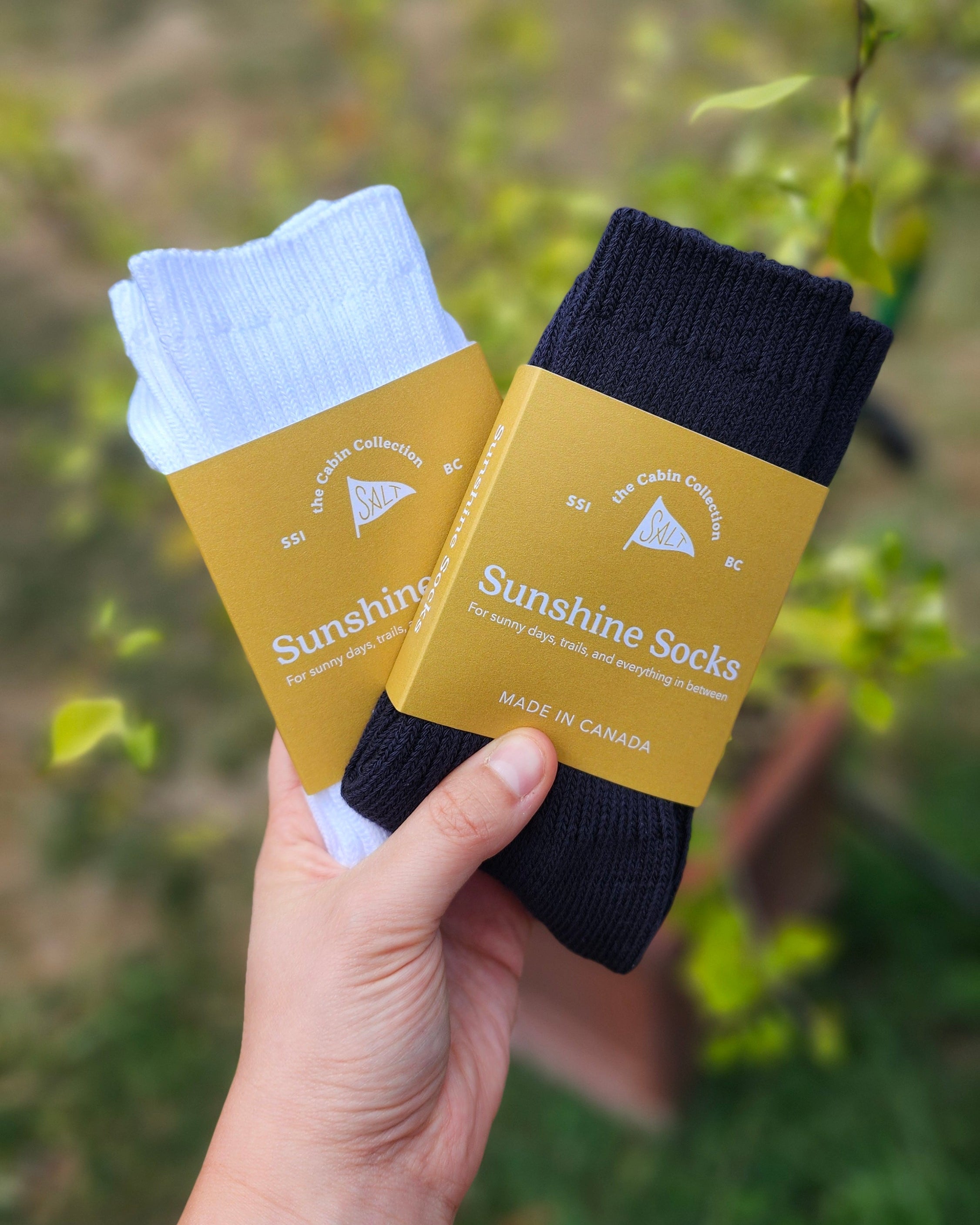 A hand holding two packaged socks against a blurred outdoor background of green foliage. One sock is navy and the other is white, both wrapped in yellow paper bands with the brand name Sunshine Socks visible.